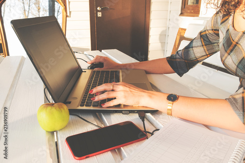 Woman's hands on laptop keyboard. Mobile smartphone, notebook and green apple on white wooden table. Working from home. Freelance, distance education concept. Selective focus on hands. 