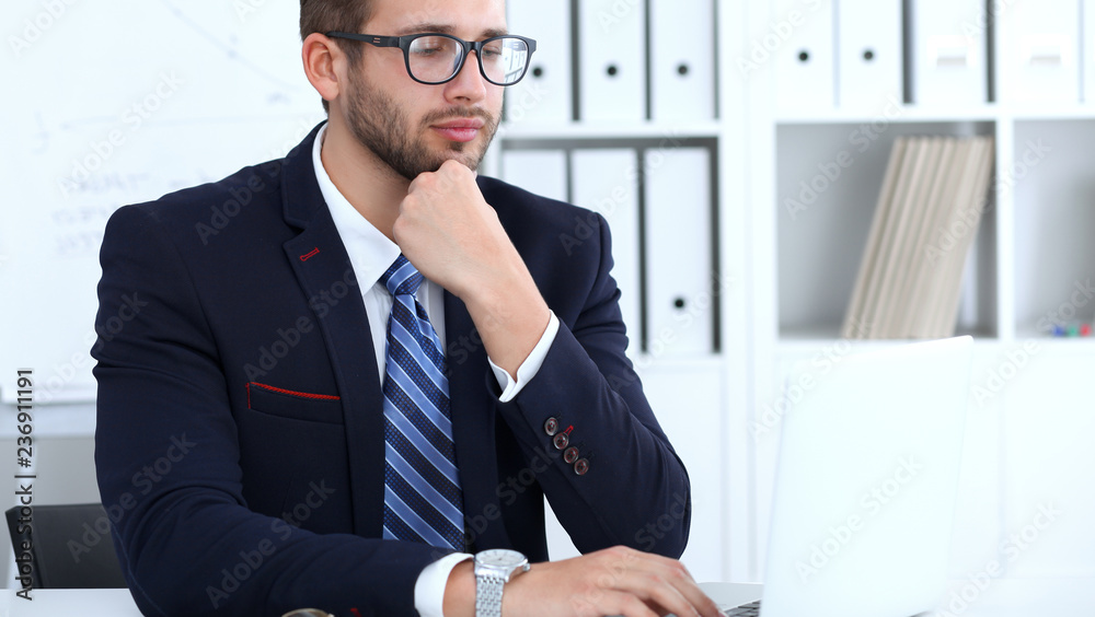 Businessman using laptop computer while sitting at the desk in office. Focus at cheerful smiling bearded man wearing glasses. Work place and success concept Businessman using laptop computer while sitting at the desk in office. Focus at cheerful smiling bearded man wearing glasses. Work place and success concept