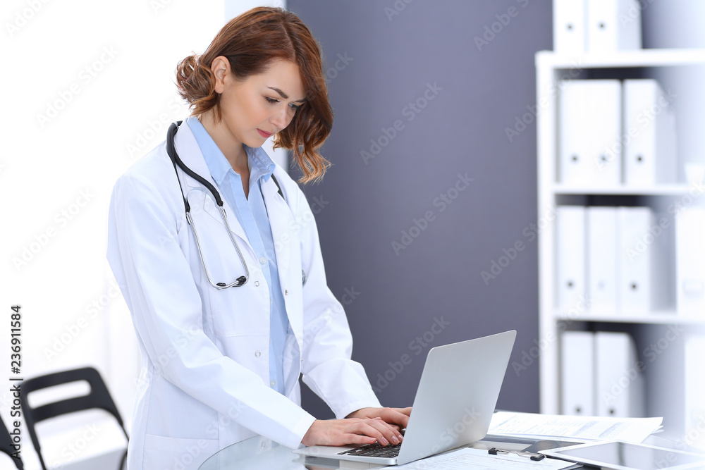Happy doctor woman at work. Portrait of female physician using laptop computer while standing near reception desk at clinic or emergency hospital. Medicine concept Happy doctor woman at work. Portrait of female physician using laptop computer while standing near reception desk at clinic or emergency hospital. Medicine concept