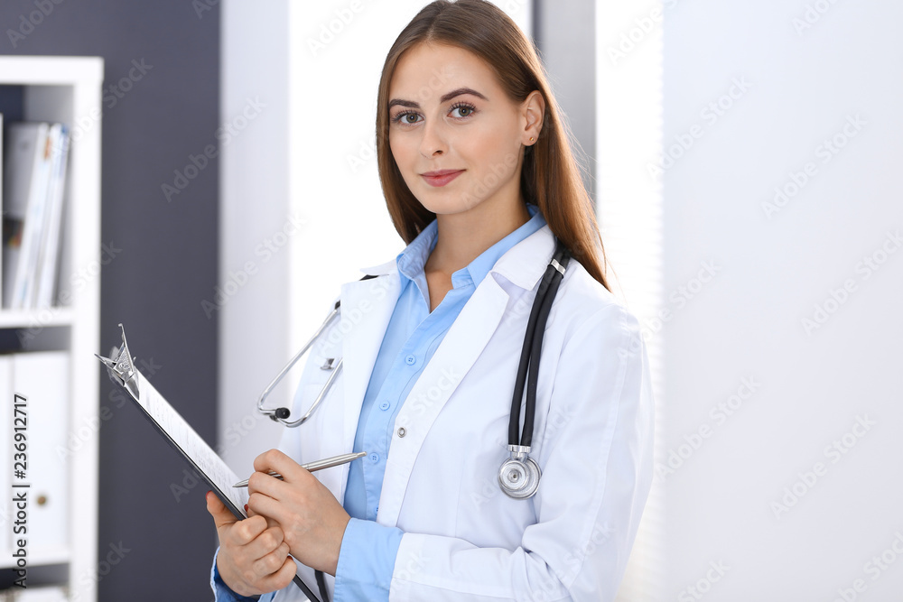 Doctor woman filling up medical form while standing near window in hospital office. Happy physician at work. Medicine and health care concept Doctor woman filling up medical form while standing near window in hospital office. Happy physician at work. Medicine and health care concept