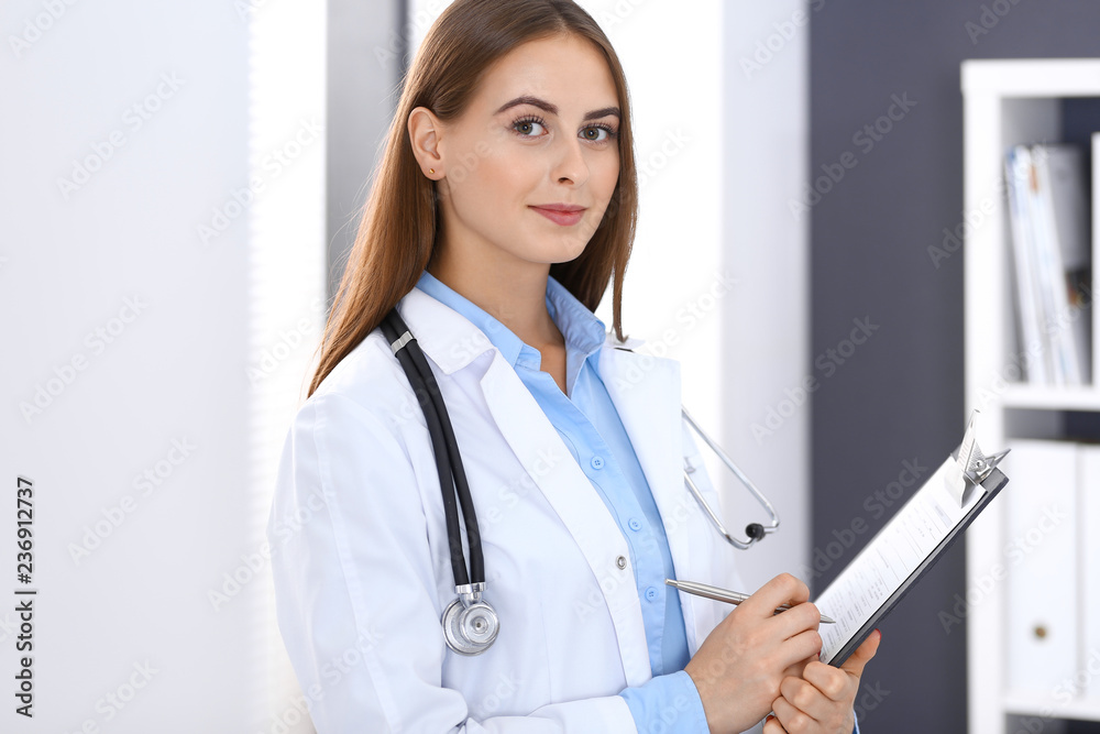 Doctor woman filling up medical form while standing near window in hospital office. Happy physician at work. Medicine and health care concept Doctor woman filling up medical form while standing near window in hospital office. Happy physician at work. Medicine and health care concept