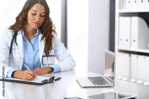 Unknown doctor woman filling up medical form while sitting at the desk in hospital office. Physician at work. Medicine and health care concept