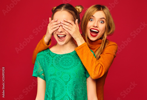 Photo of two beautiful blond ladies standing isolated over red background. Smiling girl covering eyes with hands to her friend. Positive woman models hugging