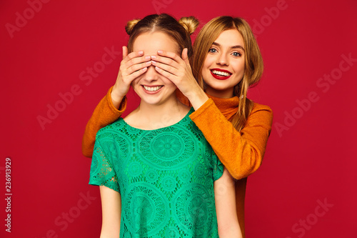 Photo of two beautiful blond ladies standing isolated over red background. Smiling girl covering eyes with hands to her friend. Positive woman models hugging