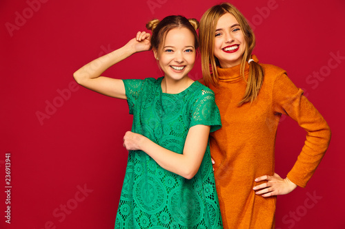 Two young beautiful blond smiling hipster girls posing in trendy summer clothes. Carefree women isolated on red background. Positive models going crazy and hugging