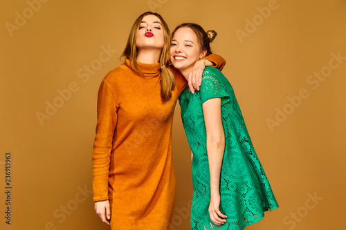 Two young beautiful blond smiling hipster girls posing in trendy summer clothes. Carefree women isolated on golden background. Positive models going crazy and hugging