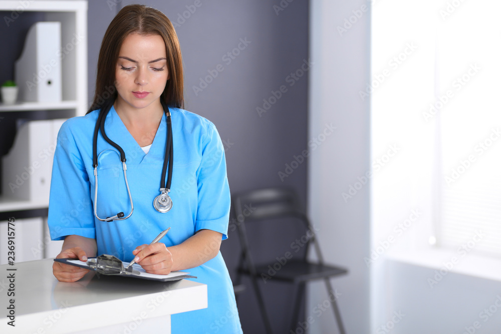 Doctor woman at work. Portrait of female physician filling up medical form while standing near reception desk at clinic or emergency hospital. Medicine and healthcare concept Doctor woman at work. Portrait of female physician filling up medical form while standing near reception desk at clinic or emergency hospital. Medicine and healthcare concept