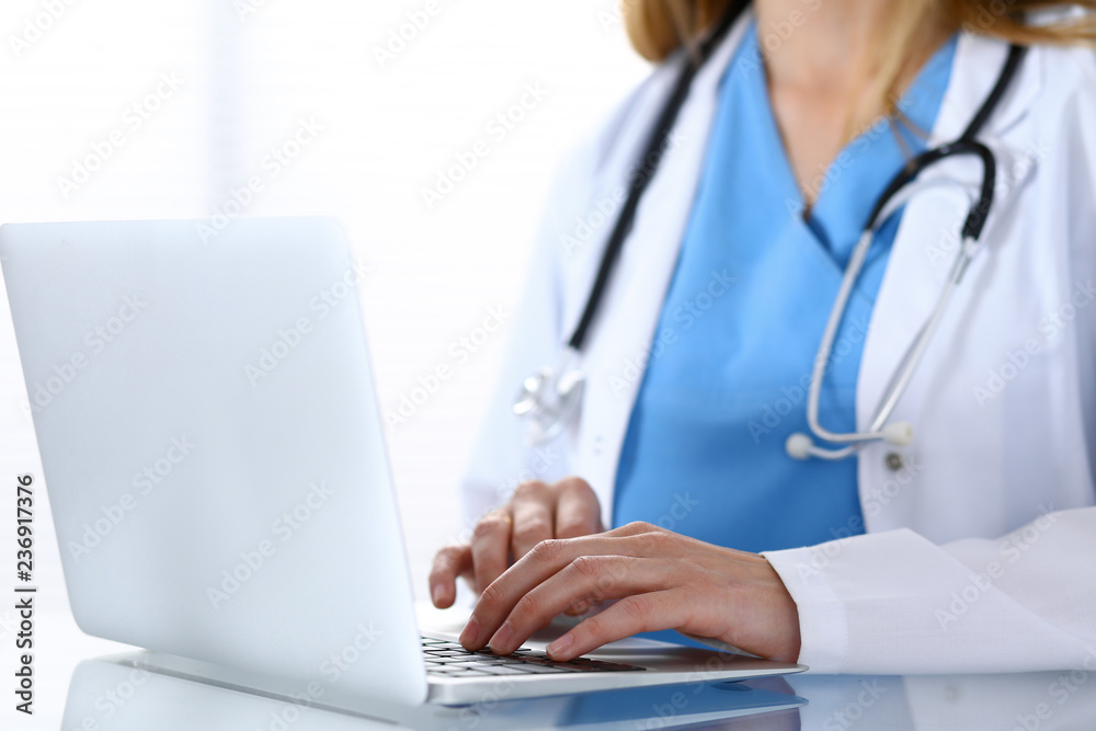 Doctor typing on laptop computer while sitting at the glass desk in hospital office. Physician at work. Medicine and healthcare concept Doctor typing on laptop computer while sitting at the glass desk in hospital office. Physician at work. Medicine and healthcare concept
