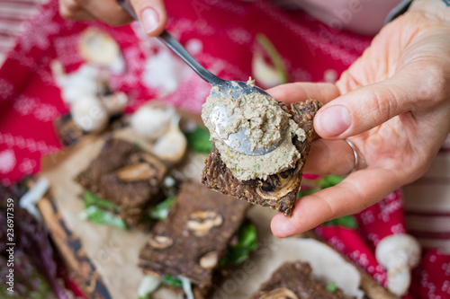Crispy rye and flax seeds breads dehydrated and stuffed with fresh green salad leaves, mushrooms. Woman hands spread hummus paste. Vegan, vegetarian snack on Christmas red tablecloth background