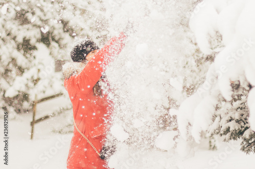 Young woman in red down jacket and black hat having fun by shaking a tree with a lot of snow falling down while is also snowing