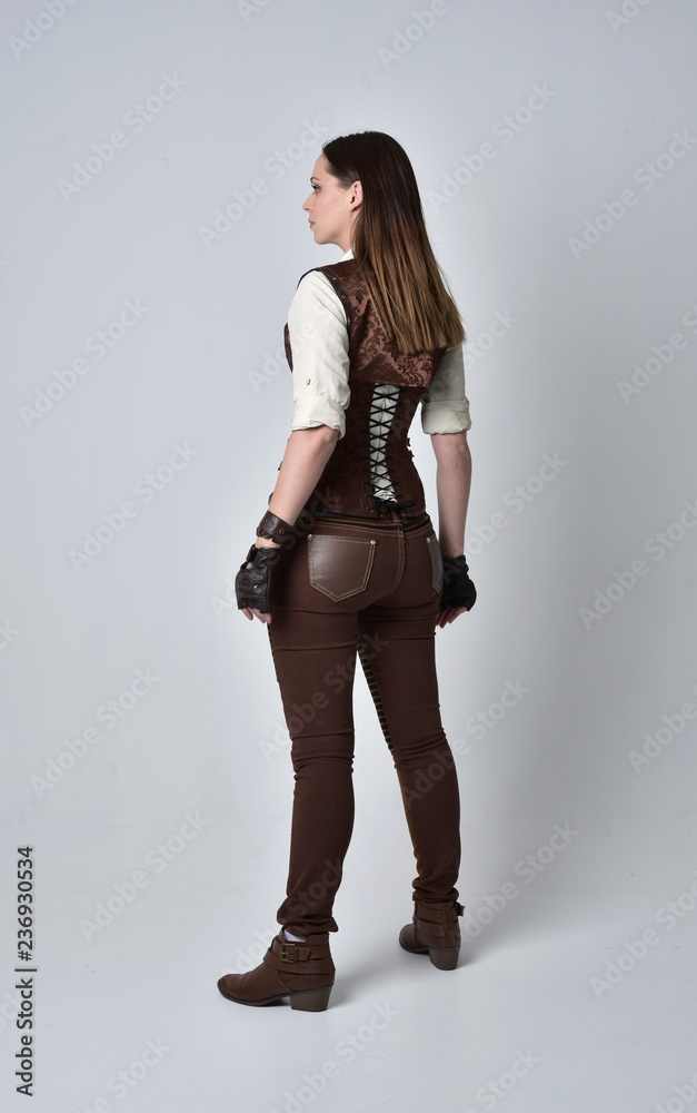 full length portrait of brunette girl wearing brown leather steampunk outfit. standing pose with back to the camera, on grey studio background. full length portrait of brunette girl wearing brown leather steampunk outfit. standing pose with back to the camera, on grey studio background.