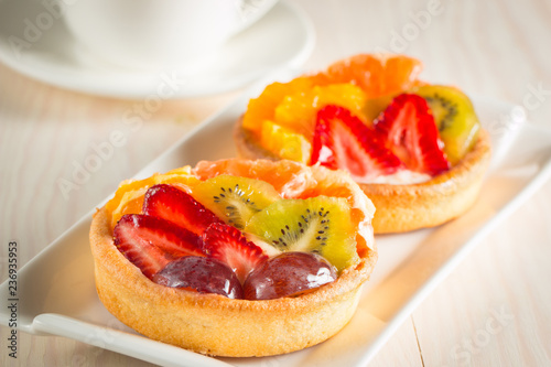 Photo of a fruit and berry tart dessert with toss sugar on wooden background. Fresh delicious sweet cake with raspberries, grapes, strawberries, cherry, kiwi, grapefruit and cream.