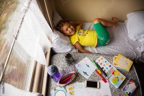 Little girl travelling by train, laying on the fisrt shelf and looking up, top view on the table with travel needs photo