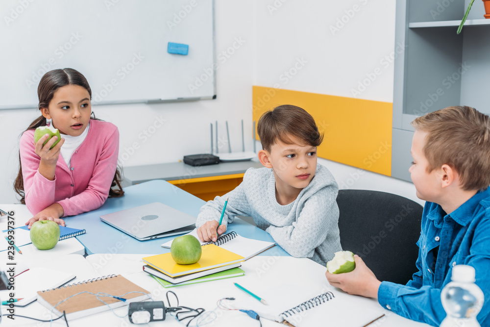 classmates sitting at table, having break, talking and eating apples in classroom classmates sitting at table, having break, talking and eating apples in classroom