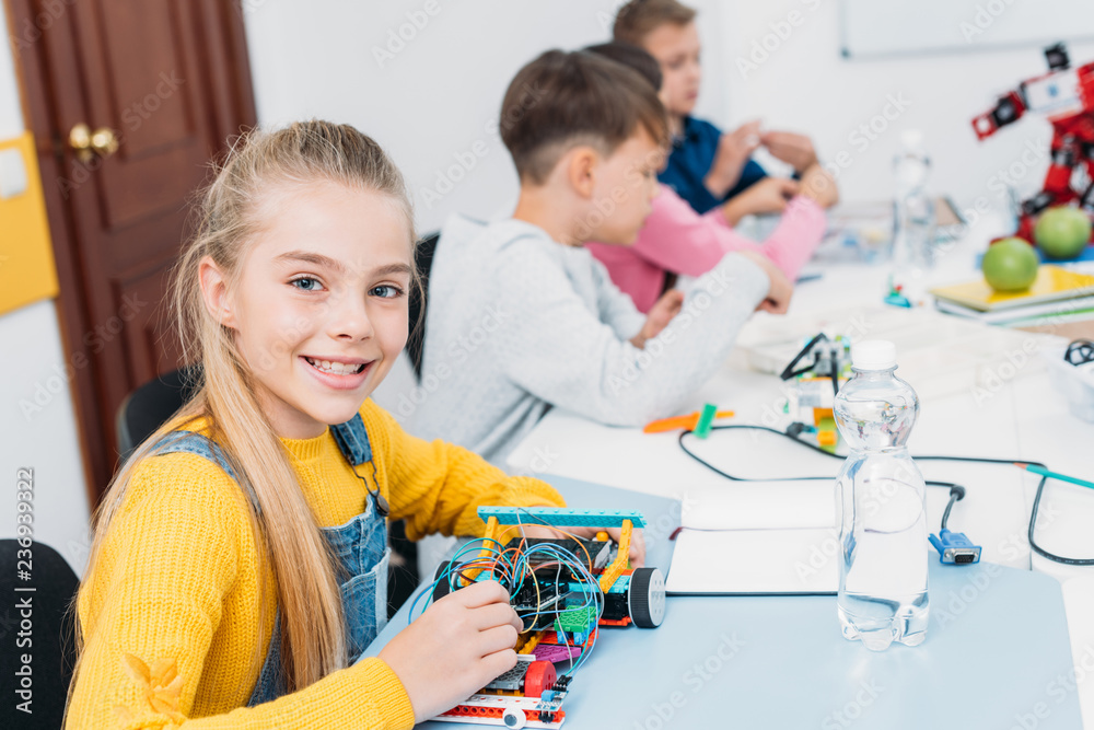 adorable schoolgirl holding robot model, looking at camera while classmates having STEM lesson adorable schoolgirl holding robot model, looking at camera while classmates having STEM lesson