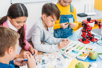 schoolchildren making red robot with details and electric kit at desk in stem education class