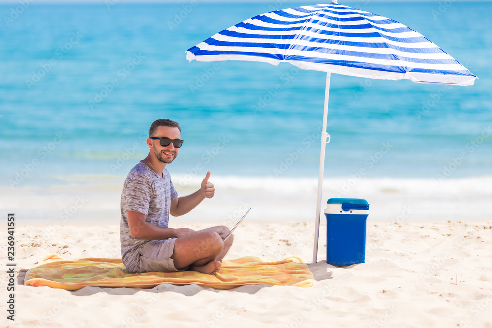 Happy young man in the beach under umbrella near the ocean working on his laptop and showing thumbs up Happy young man in the beach under umbrella near the ocean working on his laptop and showing thumbs up