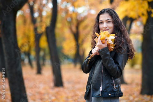 Pretty woman is posing with bunch of maple s leaves in autumn park. Beautiful landscape at fall season.
