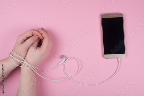 Hands tied with a cord of earbuds and a mobile phone on a pastel pink background (top view) as the mobile or cell phone addiction concept photo