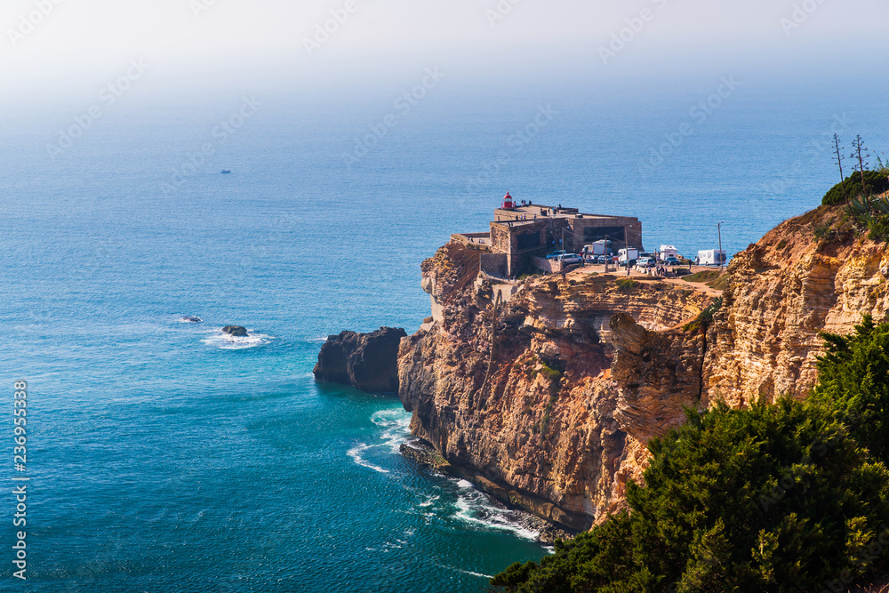 Ancient fort at seaside of Portugal, Nazare. Lighthouse on the rocky cliff. Atlantic ocean coast shoreline. Slopes of promontory are covered with green vegetation. Waves crashing on the rocks