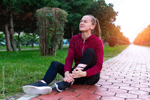 A young, fitness girl in sneakers and sportswear was injured while jogging and sports training. Leg injury and muscle strain, and ankle sprain accident during sports exercise.