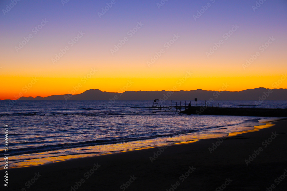 The beach and the pier at sea after sunset, a beautiful orange sky next to the silhouettes of the mountains.