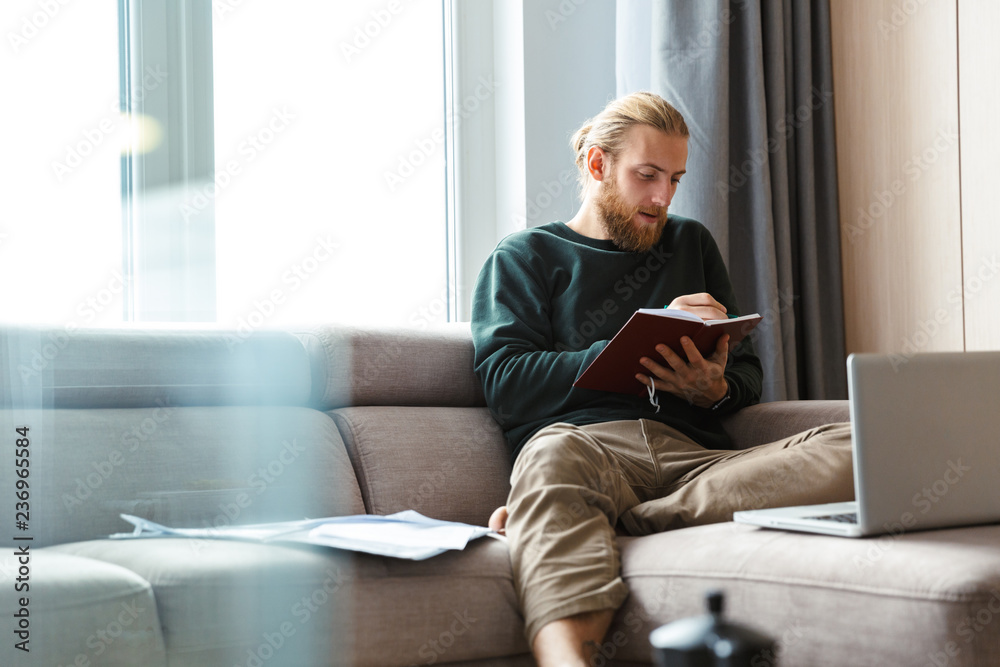 Concentrated young bearded man sitting in home writing notes. Concentrated young bearded man sitting in home writing notes.