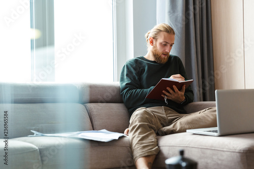 Concentrated young bearded man sitting in home writing notes.