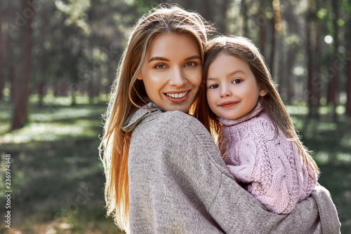 Happy, stylish little curly girl is hugging her beautiful mother in forest