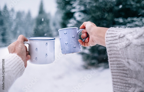 Man and woman hands in knitting mittens taking cups of hot drink. Sunny winter forest glade landscape on background. Winter forest concept image.