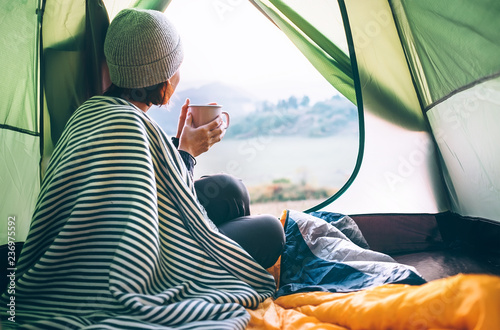 Young woman covered with warm plaid meets cold morning in sitting in a touristic camping tent with a cup of hot tea. Romantic camp traveling concept.