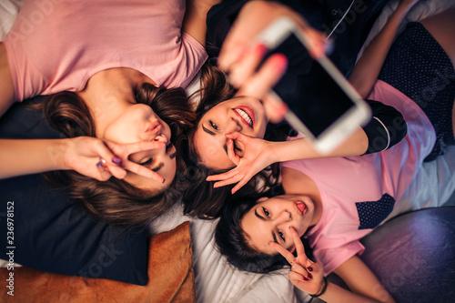 Three playful young women lying on bed. They pose on camera and make different poses. Model in middle hold white phone in hand.