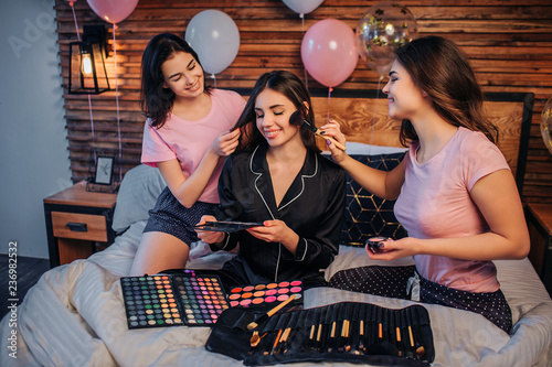 Three happy young women sit on bed in festive room. Two girls do haircare and makeup for third one. There are brushes and shadown on bed. Model in black pajamas hold one set of shadows in hands. photo