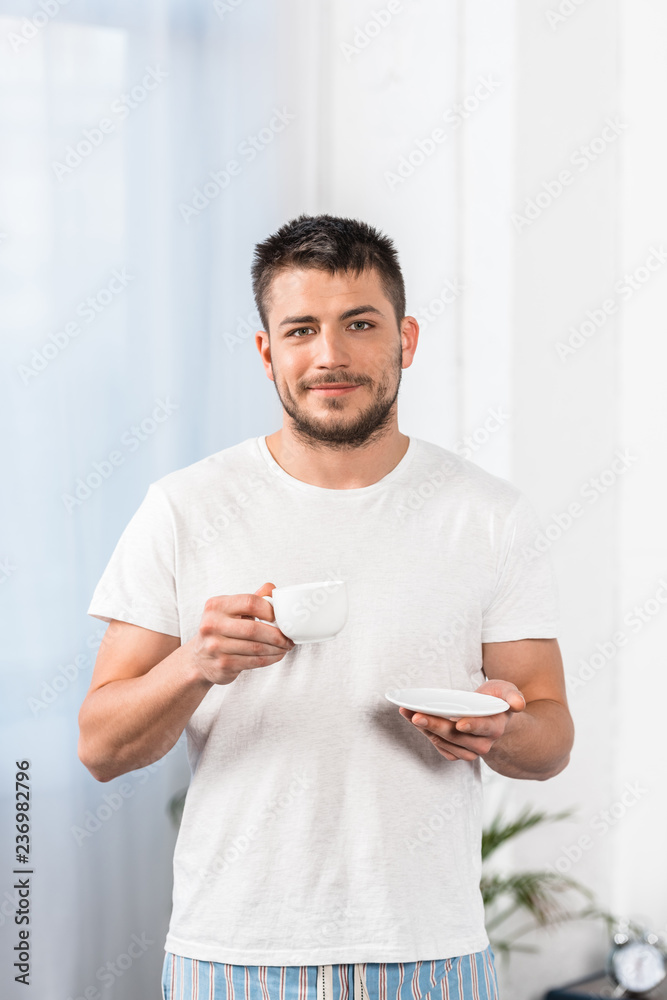 handsome man in shirt and pajamas holding cup of coffee and looking at camera in morning in bedroom handsome man in shirt and pajamas holding cup of coffee and looking at camera in morning in bedroom