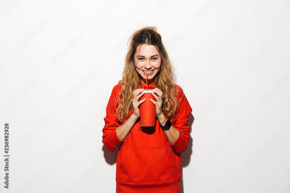 Fototapeta premium Image of attractive woman 20s wearing red sweatshirt drinking soda from paper cup using straw, isolated over white background