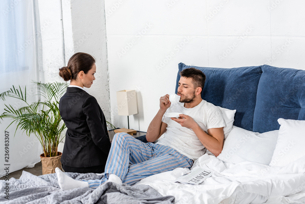 Obraz premium girlfriend in suit sitting on bed near boyfriend in pajamas drinking coffee in morning at weekday, gender stereotypes concept