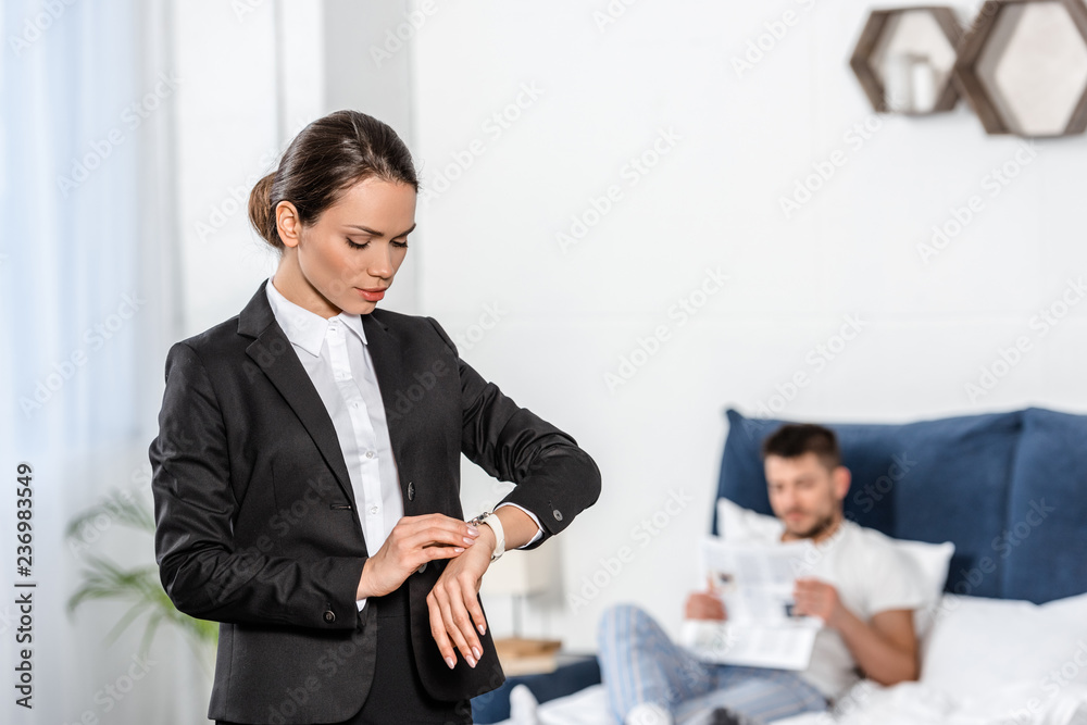 girlfriend in suit checking time at wristwatch and boyfriend in pajamas reading newspaper in bedroom in morning, gender equality concept girlfriend in suit checking time at wristwatch and boyfriend in pajamas reading newspaper in bedroom in morning, gender equality concept