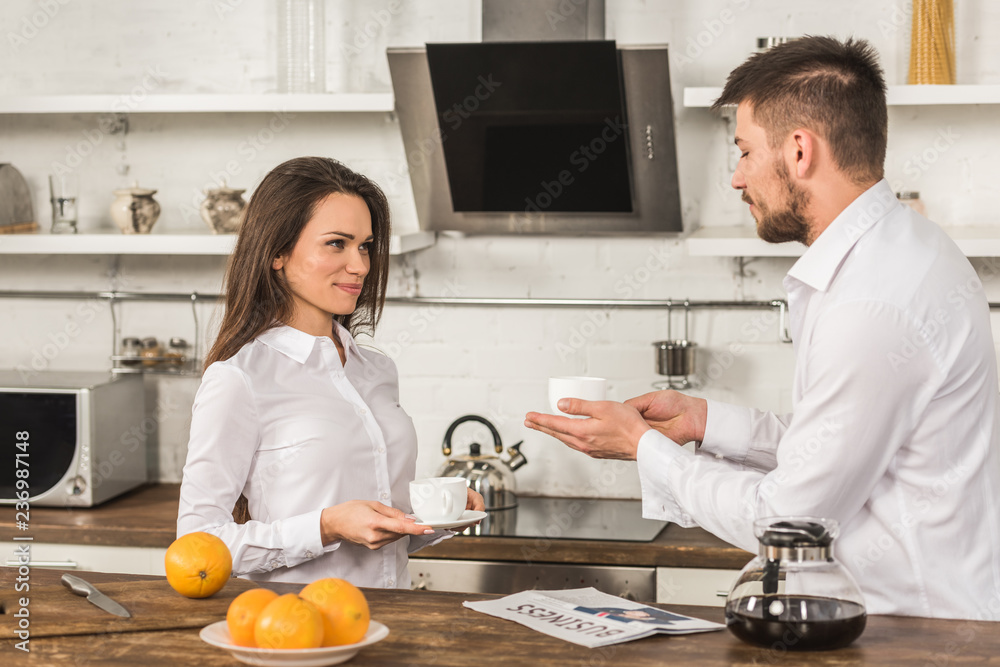 cheerful boyfriend and girlfriend in shirts standing with cups of coffee in morning at kitchen, gender equality concept cheerful boyfriend and girlfriend in shirts standing with cups of coffee in morning at kitchen, gender equality concept