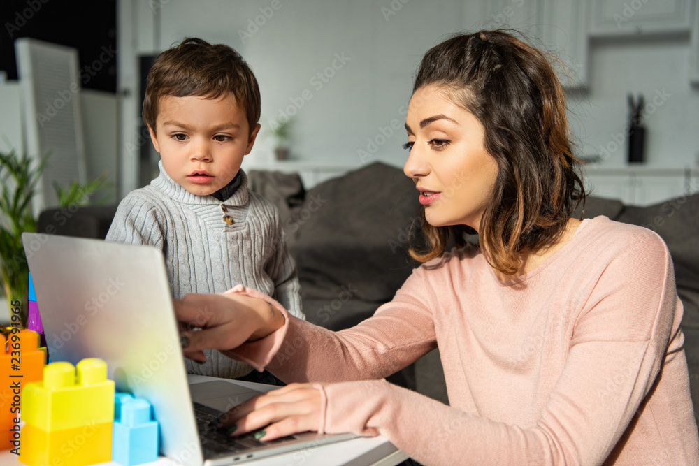 attractive woman pointing at laptop screen to her little son at home attractive woman pointing at laptop screen to her little son at home