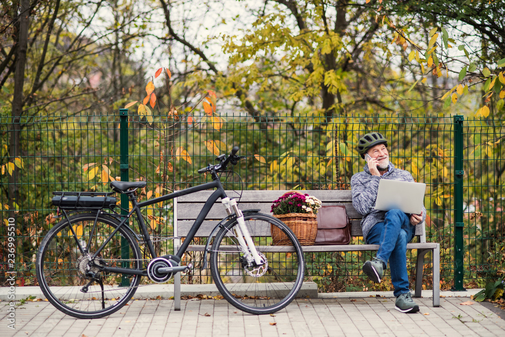Obraz premium Senior man with electrobike sitting on a bench outdoors in town, using laptop and smartphone.