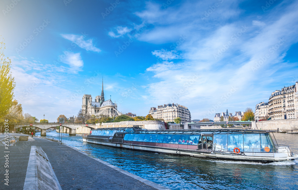 Fototapeta premium Cathedral of Notre Dame de Paris at sunny autumn afternoon. Embankment of the Seine. Boat on scenic route. Tourists a walk and relax in warm weather. Blurred unrecognizable faces. Paris. France.