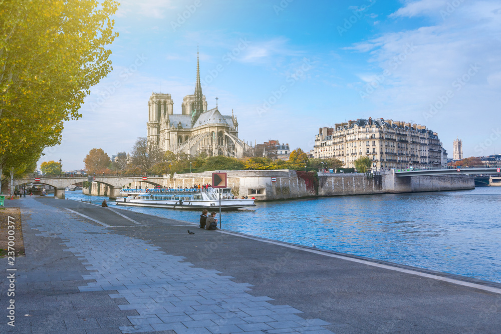 Obraz premium Cathedral of Notre Dame de Paris at sunny autumn afternoon. Embankment of the Seine. Boat on scenic route. Tourists a walk and relax in warm weather. Blurred unrecognizable faces. Paris. France.