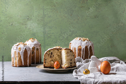 Set of traditional Russian and Ukrainian Easter cake Kulich Paska bread glazed with almond, whole and sliced, served on ceramic plate with cloth over grey table with green wall as background. photo