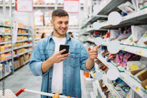 Man with cart makes purchase by the list on phone