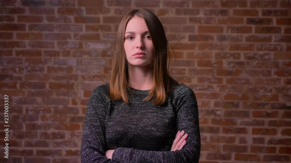 Portrait of caucasian ginger woman with serious eyes watching directly into camera on bricken background.