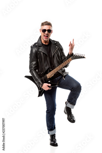 excited stylish man in leather jacket holding electric guitar and showing rock sign isolated on white