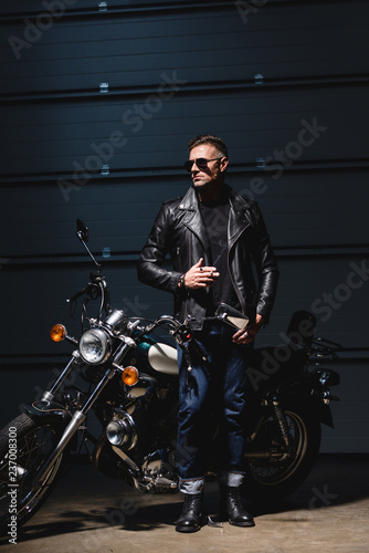 classic guy in sunglasses standing by motorcycle in garage and smoking cigarette