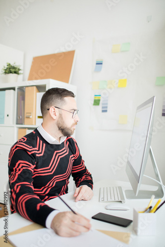 Serious concentrated handsome bearded businessman in glasses sitting at table and making note while holding online analysis, he using computer