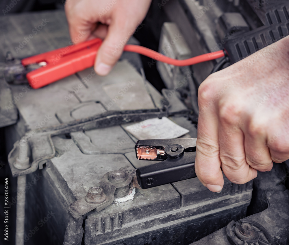 Man recharging a dead car battery in winter time using start-charger. Close up hands of a man with two contact clips from battery charger for vehicles. People under bonnet with starter cables