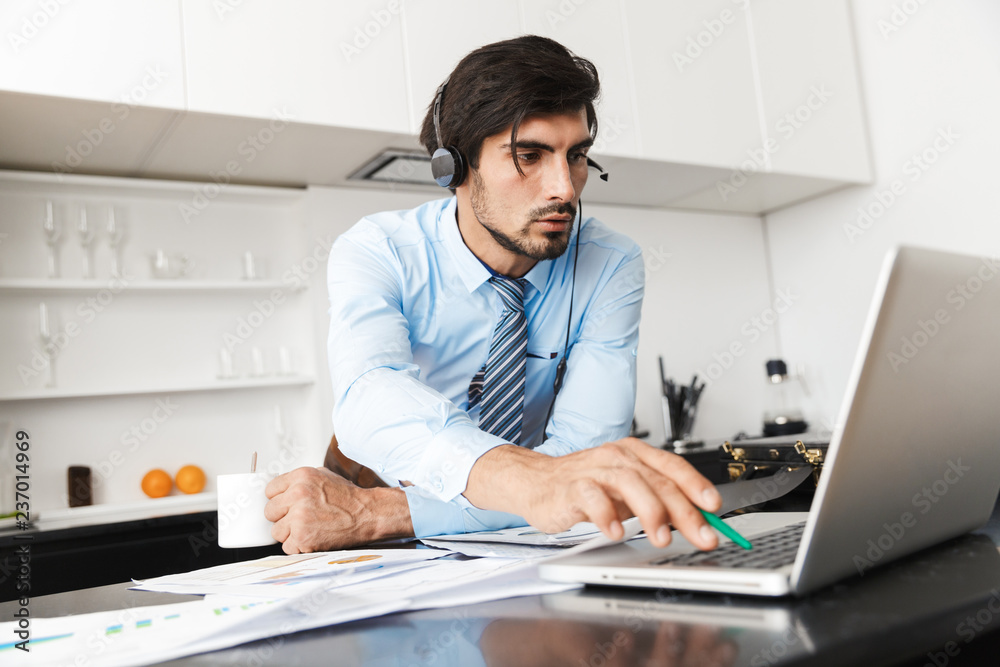 Concentrated young business man at the kitchen wearing headphones work with documents using laptop computer. Concentrated young business man at the kitchen wearing headphones work with documents using laptop computer.